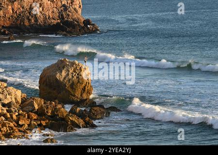 Man fishing from a large boulder off Praia do Zavial in the North Atlantic, in the Algarve region of Portugal; Raposeira, Faro, Portugal Stock Photo