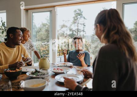 Happy boy talking while having lunch with family members on dining table at home Stock Photo