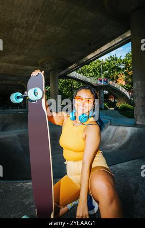 Portrait of smiling young woman crouching with longboard under bridge Stock Photo