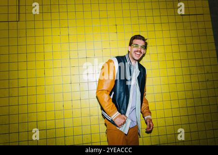 Portrait of happy young man in varsity jacket standing near yellow wall Stock Photo