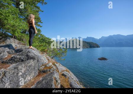 Woman standing on rocks looking at the British Columbia mainland from an island on a warm sunny day; Vancouver, British Columbia, Canada Stock Photo