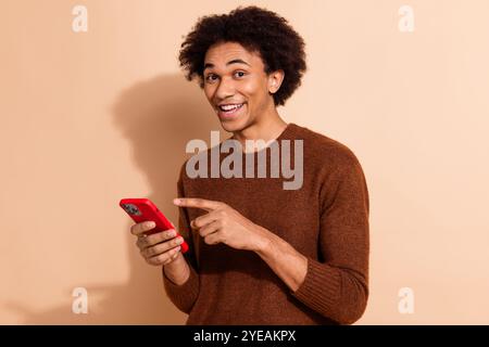 Young man pointing at smartphone screen excitedly on beige background, wearing casual sweater conveying cheerful and shopping concepts Stock Photo