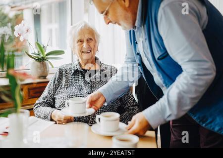 Elderly man serving tea to smiling senior man sitting at retirement home Stock Photo