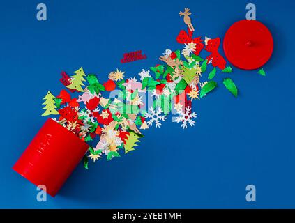 Colorful Christmas glitter confetti mix falling from red Xmas cookies tin decorated with classic bells on blue background. Minimal Christmas concept. Stock Photo