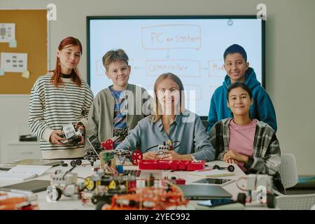 Portrait of smiling teacher sitting at table and surrounded by multiracial students in classroom of robotics with huge number of robots and its parts Stock Photo