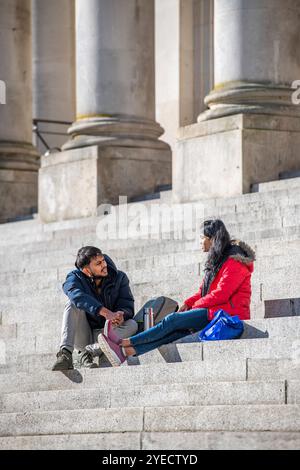 young Asian couple sitting together on the steps of Portsmouth Guildhall chatting on a bright sunny day Stock Photo