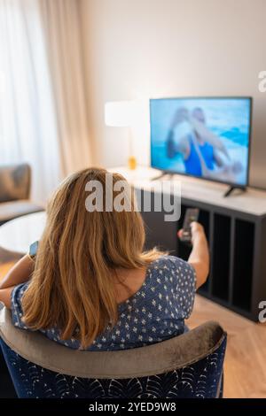 Woman sitting in an armchair with her back to her and changing the TV channel with the remote control Stock Photo