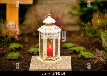 a white vintage lantern with a red burning candle stands on a marble slab on a grave with a cross in the blurred background Stock Photo