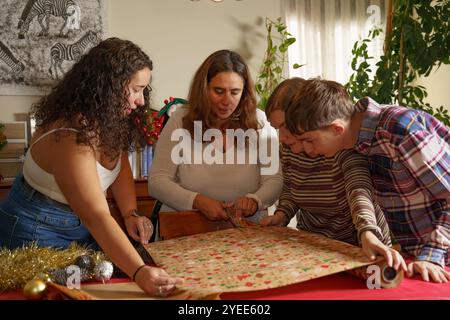 A group of four people, including two women and two men, are gathered around a table, engaged in wrapping gifts. They are using festive wrapping paper Stock Photo