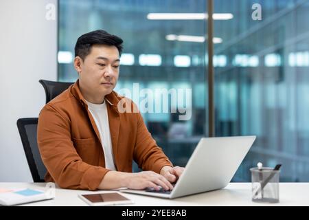Asian business man focused on laptop work in modern office. Professional in casual wear embraces digital technology and innovation in office setting. Nearby is a smartphone and notepad. Stock Photo