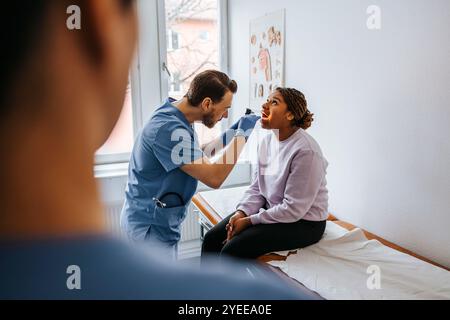 Male doctor examining throat of woman using tongue depressor and flashlight in examination room Stock Photo