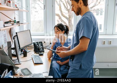 Female nurse with hand on chin sitting next to male colleague writing in clinic Stock Photo