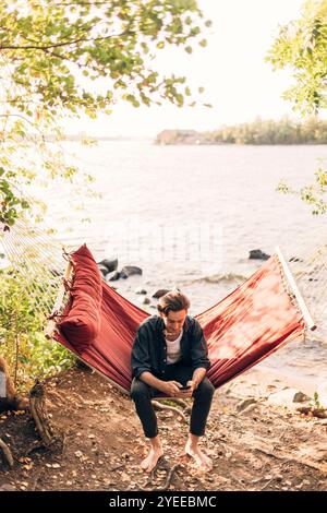Full length of man using smart phone while sitting in hammock near lake Stock Photo