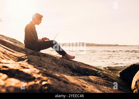 Side view of young man using laptop while sitting on rock near lake at sunset Stock Photo