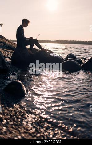 Side view of young man using laptop while sitting on rock by lake at sunset Stock Photo