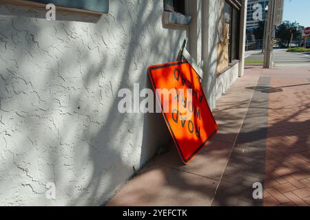 Orange Road Work Ahead sign on brick sidewalk leaning up beside a gray building. Leading lines down the street towards intersection and blue sky. Stock Photo