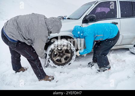 Travel in wintertime, pair of unrecognizable friends, man and woman, putting chains on the wheels of an SUV vehicle, in order to be able to move forwa Stock Photo