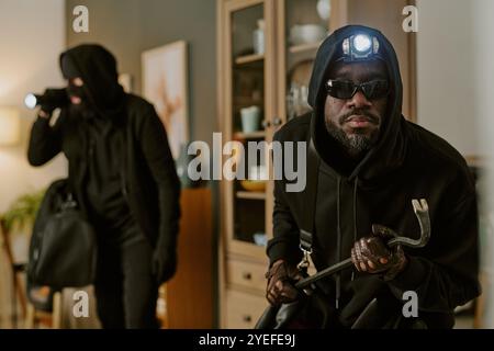 Medium close up of African American thieve with headlamp and crowbar in hands looking at camera during trespassing Stock Photo