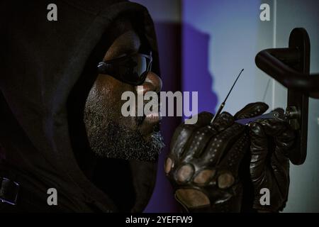 Medium close up of African American thief hiding his head under hood holding lockpick while trying to open door Stock Photo