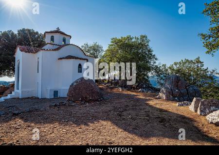 A quaint white chapel stands on rocky terrain in Maniaki, Greece, surrounded by trees and sunlight, reflecting traditional Greek architecture and spir Stock Photo