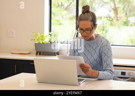 Mature woman reading documents at home using laptop, concentrating on work Stock Photo