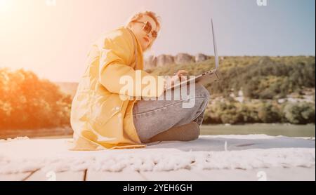 Man Laptop Outdoor Work - A man sits on a rooftop, wearing sunglasses, and works on a laptop with a sunny view. Stock Photo