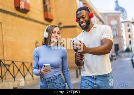 Multi-ethnic couple using mobile phone and headphones in the city sharing content and smiling Stock Photo