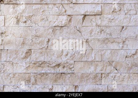 Stone wall made of beige tiles arranged in a staggered pattern. The tiles have a rough texture and visible cracks, giving the wall a rustic appearance. Stock Photo