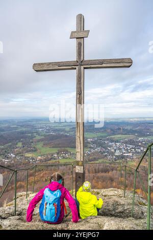 Two hikers rest by the wooden summit cross on Oresnik granite rock formation, taking in the panoramic views of Hejnice and the Jizera Mountains in Czechia. Stock Photo