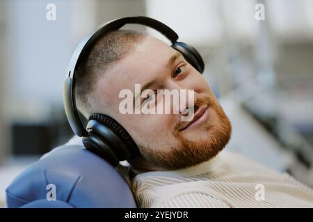 Man with headphones sitting in a relaxed position while enjoying music and smiling. He is looking straight at the camera with a friendly expression, seated indoors Stock Photo
