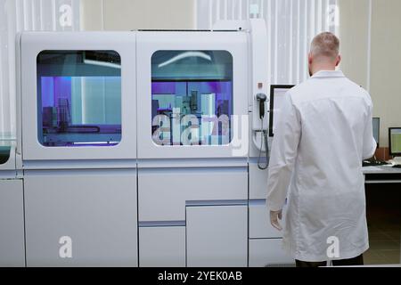 White-coated scientist operating modern laboratory equipment, immersed in task, in a high-tech setting featuring advanced machinery and screens, focusing on materials handling Stock Photo