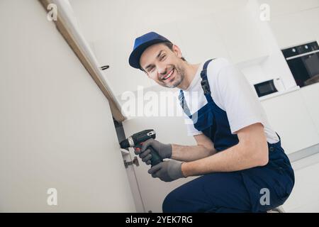 Smiling handyman in blue overalls using a cordless drill for home renovation in a modern kitchen setting Stock Photo
