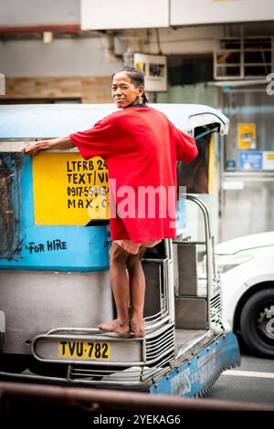 A young Filipino many holds on to the back of a jeepney bus speeding through Manila City, The Philippines. Stock Photo