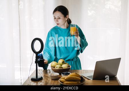 Young woman creating social media content on healthy lifestyle with fruits and juice in kitchen. Wellness influencer and food vlogging concept Stock Photo