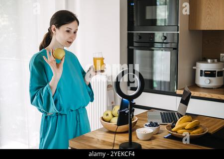 Young woman creating social media content on healthy lifestyle with fruits and juice in kitchen. Wellness influencer and food vlogging concept Stock Photo