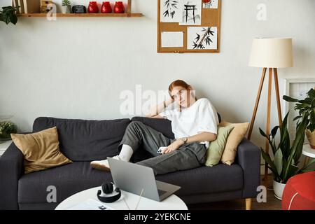 A young man in casual attire sits comfortably on a couch, working on a laptop in a bright space. Stock Photo