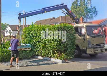 Back view male truck delivery driver operating hand held controls of lorry mounted offload crane delivering pallet of bagged cobbles Essex England UK Stock Photo