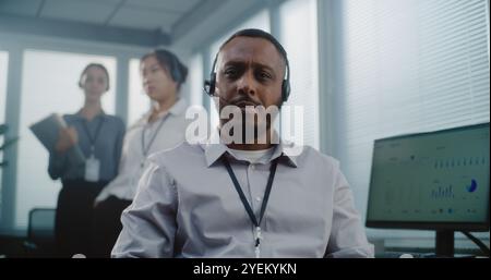 Call center office: Close up portrait of friendly African American technical customer support specialist wearing headset sitting, looking at camera. Team of hotline operators standing in background. Stock Photo