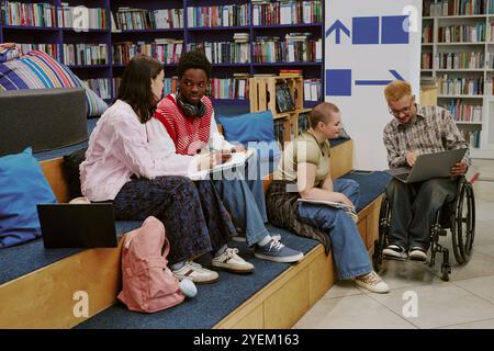 Friends sitting together, collaborating on project using laptop and notes in university library. Bookshelves filled with resources in bright, modern study area Stock Photo