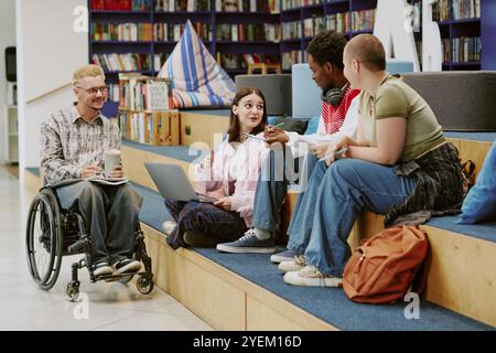 Group of diverse students engaging in an animated study session while sitting on steps of a modern library setting, surrounded by books and technology Stock Photo