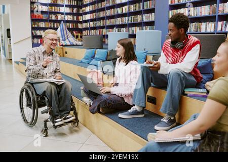 Group of students interacting in library, discussing ideas while using laptops. One student in wheelchair engaged with peers, creating inclusive learning environment Stock Photo