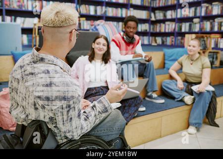 Group of diverse students sitting in modern library engaged in study session on tiered seating. One student in wheelchair, all focused and interacting, surrounded by shelves of books Stock Photo