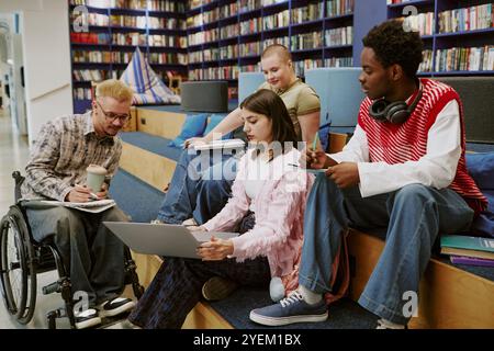 Group of students collaborating in modern library setting, with one student in wheelchair holding a coffee, others engaged with books and laptop Stock Photo