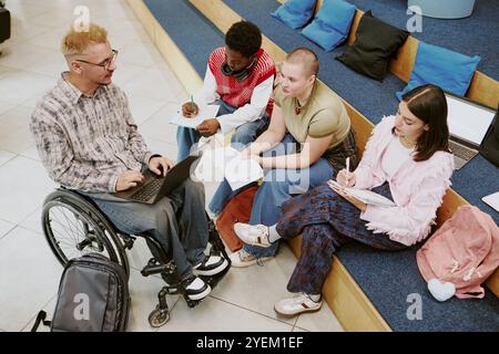Group of diverse university students engaging in studying, sitting on campus steps, with notes and laptops, involving student in wheelchair, promoting collaborative learning atmosphere Stock Photo