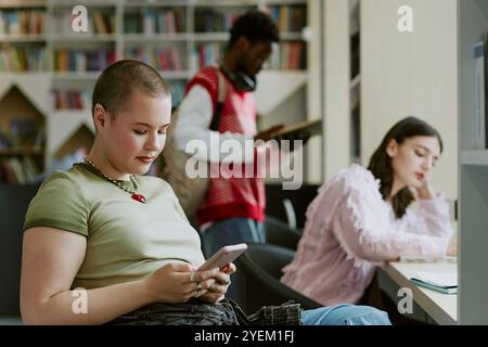 Students studying in a library setting with bookshelves in background, using smartphones and reading material. Scene captures focus and concentration while engaging with digital and printed resources Stock Photo