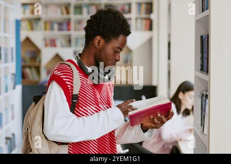 Young man with backpack reading book while wearing headphones standing in modern library setting with bookshelf background Stock Photo