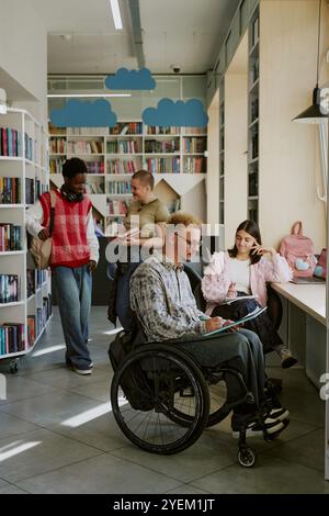 Group of students studying in a modern library with cloud-themed decor, with one student in a wheelchair working at desk and others interacting in background Stock Photo
