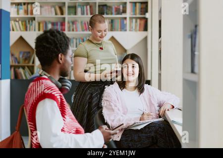 Group of diverse students studying in a modern library, engaging in conversation and taking notes. Shelves filled with books in the background creating an academic atmosphere Stock Photo