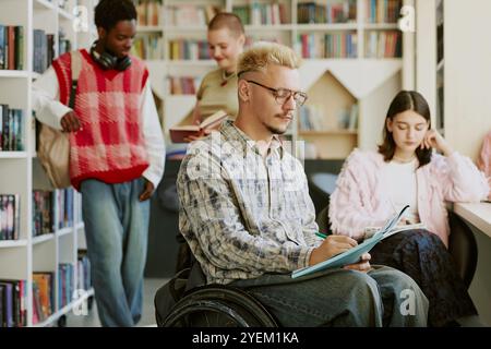 Group of diverse students engaging in collaborative study session in cozy library setting, with one student using a laptop and another in a wheelchair, deep in discussion Stock Photo