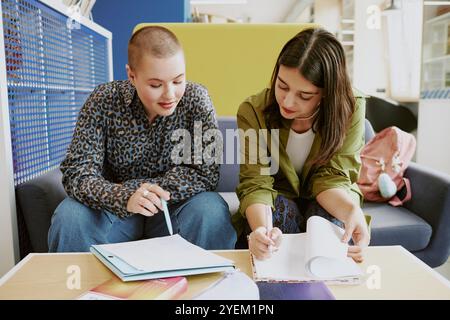 Two young women collaborating at work reviewing documents in modern office. One woman with short hair in leopard print blouse and other with long hair in green jacket Stock Photo